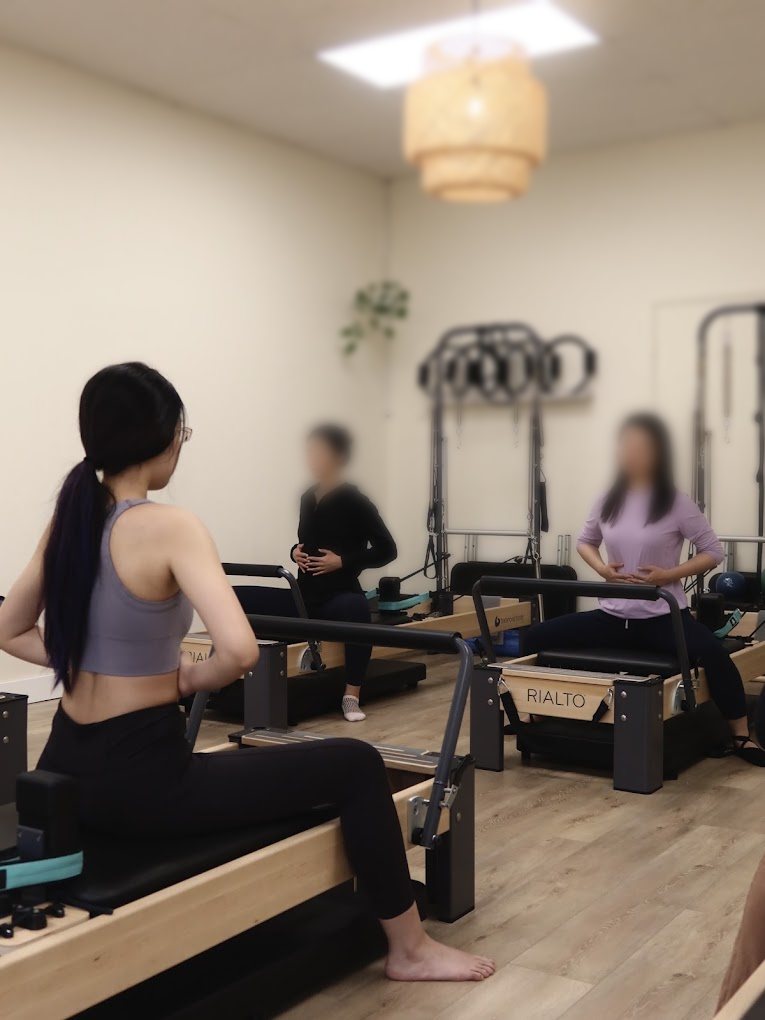 A reformer class in progress at Aloe Pilates, clients seated on Balanced Body RIALTO reformers under a woven pendant light